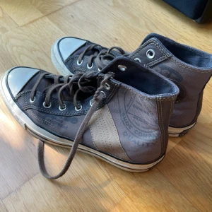 Converse All Star bruna high-top sneakers - Säljer ett par Converse All Star high-top sneakers i brunt tyg med beige detaljer och klassisk vit gummitå. Skorna har snörning, rund tå och den ikoniska Converse-loggan på sidan. Perfekta för dig som gillar en avslappnad och cool stil.