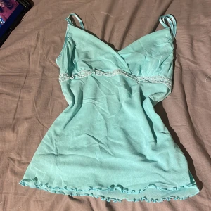 Turkos linnetopp från Etam med spets - Supersöt turkost linnetopp från Etam med tunna axelband, v-ringning och dekorativ spets med pärldetaljer under bysten. Toppen har volangkant nertill och är gjord i ett lätt, luftigt material. Perfekt för en fräsch och somrig look! Sitter som en S/xs pris går och diskuterar 