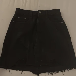 Svart jeanskjol med fransig kant - Svart kort jeanskjol med rå, fransig nederkant och klassiska femficksdetaljer. Kjolen har bälteshällor och knappstängning framtill. Perfekt för en avslappnad och cool stil.