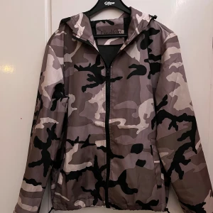 Camo windbreaker  - Säljer en snygg vindjacka från Lindfors med grått, beige och svart camouflage-mönster. Jackan har huva, dragkedja framtill och justerbar nederkant med snörning. Perfekt för dig som vill sticka ut. Passar perfekt till hösten 