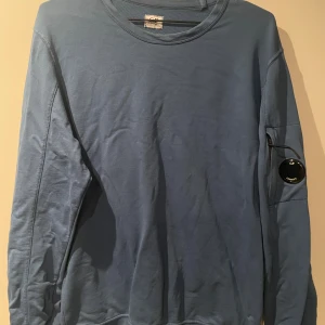 Blå crewneck från C.P. Company - Säljer en blå långärmad crewneck från C.P. Company med deras ikoniska linsdetalj på ärmen. Tröjan är i mjuk bomull och har en clean look med rund hals. Perfekt för dig som gillar streetwear och vill ha något stilrent men ändå unikt. Skick 9/10 med nytt märke, pris går ABSOLUT att diskuteras ( tar även emot byten) 🤩