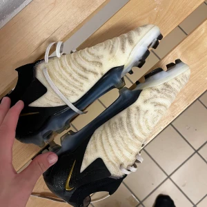 Nike fotbollsskor svart/vit/guld - Säljer ett par Nike fotbollsskor med svart och vit ovandel, guldigt Swoosh på sidan och vita snören. Skorna har mönstrad textil och syntetmaterial, samt dobbar för spel på gräs. Snygg design med tydliga detaljer och modern look.