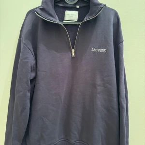 Mörkblå half zip tröja Les Deux - Säljer en mörkblå långärmad tröja från Les Deux med halv dragkedja och hög krage. Tröjan är i bomull och har en clean look med broderad logga på bröstet. Perfekt för dig som gillar stilrena och bekväma plagg. 
