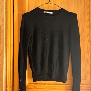 Svart stickad tröja från Zara med knappar - Svart stickad tröja från Zara i storlek S. Tröjan har lång ärm och coola guldfärgade knappar vid ärmsluten som ger en snygg detalj. Enkel och stilren modell med rund hals, perfekt till jeans eller kjol.
