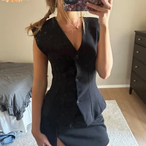 Svart figursydd topp med knappar - Snygg svart topp med markerad midja och v-ringning. Västen har tre knappar framtill och breda axlar som ger en cool siluett. Perfekt att styla över en skjorta eller t-shirt för en trendig look. Materialet är slätt och västen har en klassisk, tidlös design.