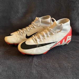 Nike Mercurial Zoom fotbollsskor - Säljer ett par Nike Mercurial Zoom fotbollsskor i vitt med svarta detaljer och röd sula. Skorna har snörning, högre skaft och är tillverkade för att ha bra grepp vilket ger dig riktigt bra grepp med bollen. Perfekta för dig som vill sticka ut med stil och prestanda. 