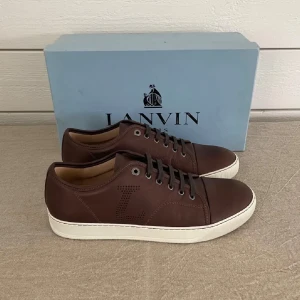 Bruna sneakers i skinn från Lanvin - Säljer ett par exklusiva sneakers från Lanvin Paris i storlek 43. Skorna är tillverkade i brunt läder med vit gummisula och har en tidlös design med perforerat L på sidan samt diskret branding på insidan. De är i mycket fint skick och levereras med originalbox. Ett par stilrena och lyxiga sneakers som passar lika bra till vardags som till mer dressade tillfällen.