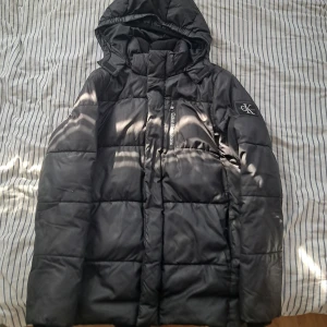 Svart pufferjacka Calvin Klein Jeans - Svart pufferjacka från Calvin Klein Jeans med huva och dragkedja framtill. Jackan har ett stilrent CK-märke på ärmen och quiltad design. Perfekt för kalla dagar och har en snygg, enkel look som funkar till det mesta.