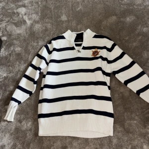 Randig tröja från U.S. Polo Assn. - Vit och marinblå randig tröja från U.S. Polo Assn. med broderad logga på bröstet. Tröjan har en halv dragkedja vid halsen, ribbade muddar och är långärmad. Storlek 50, motsvarar ungefär s skulle jag säga.