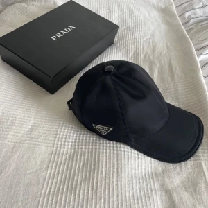 Svart keps från Prada - Snygg svart keps från Prada med klassisk böjd skärm och ikonisk metallogo på sidan. Kepsen är tillverkad i ett slätt syntetmaterial som ger en stilren och modern look. Perfekt accessoar för dig som gillar exklusiva märken och minimalistisk design. Mer info skriv pm