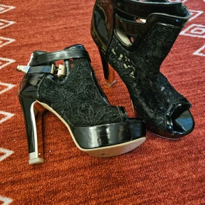 Klacksko i spets & lack ,  strl 36 - Säljer ett par grymt snygga svarta klackskor , strl 36 med hög klack och plattform. Skorna har snygga spetsdetaljer, lackade partier och öppen tå. De stängs med dragkedja bak och har ett dekorativt spänne i guld vid ankeln. Perfekta för dig som vill sticka ut. Använda ett fåtal ggr inomhus.  Älskar dem, men kan tyvärr inte längre gå i klackar . 