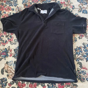 Svart frotté t-shirt från Selected Homme - Svart t-shirt från Selected Homme i storlek L, tillverkad i mjuk frotté. Klassisk krage, korta ärmar och en bröstficka ger en chill och stilren vibe. Perfekt för dig som gillar enkel men snygg design.