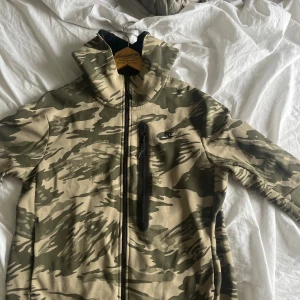Nike camo vindjacka med huva - Nike vindjacka i grön och beige camouflage med hel dragkedja och hög krage. Jackan har en bröstficka med dragkedja och huva för extra skydd. Perfekt för dig som gillar streetwear och sportig stil.