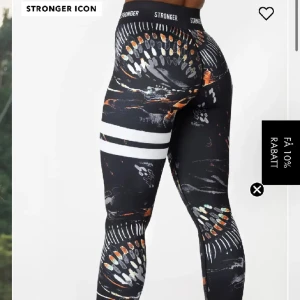 Stronger leggings med grafiskt mönster - Snygga svarta leggings från Stronger med höga vita ränder och färgstarkt grafiskt mönster i orange, vitt och grått. Tight passform och hög midja, perfekt för träning. Elastiskt material med logga i midjan för extra sportig vibe.