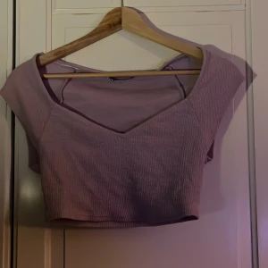 Ljuslila croppad topp från Zara - Säljer en ljuslila croppad topp från Zara med v-ringning och korta ärmar. Toppen är ribbad och har en snygg, figurnära passform som framhäver axlarna. Perfekt att matcha med höga jeans eller kjol för en trendig look.