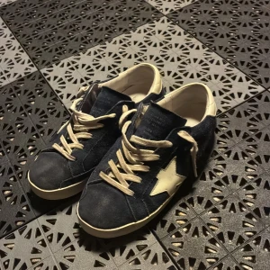 Mörkblå sneakers med vit stjärna - Snygga mörkblå Golden goose med vit stjärndetalj på sidan och vita snören. Ovandelen är i mocka och insidan är fodrad i vitt. Skorna har platt sula och klassisk rund tå. Perfekta för dig som gillar en sportig och trendig look.