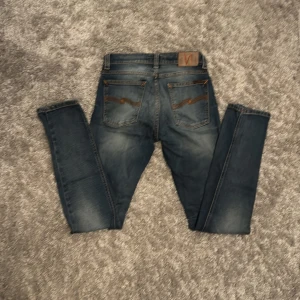 Blå skinny jeans från Nudie Jeans - Säljer ett par blå skinny jeans från Nudie Jeans i storlek 28/32. Jeansen har snygga slitningar och klassiska orangea sömmar, samt Nudie-logga på bakfickan. De är tillverkade i stretchig denim som sitter tajt och bekvämt.