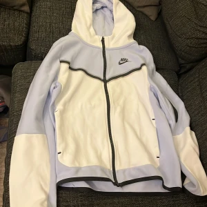 Nike hoodie i vitt och ljuslila - Nike hoodie i vitt och ljuslila med svart Nike-logga på bröstet. Tröjan har hel dragkedja, huva och svarta detaljer längs kanterna. Tillverkad i mjukt syntetmaterial, perfekt för chill eller träning. Långärmad och med snygg sportig look.