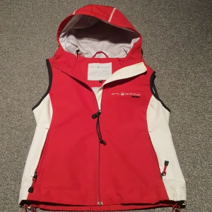 Röd Sail Racing windstopper väst - Sportig röd och vit väst från Sail Racing med huva och dragkedja framtill. Tillverkad i vindtätt material, perfekt för aktiva dagar utomhus eller på havet. Funkar också perfekt till vardagliga utflykter eller till skolan. Har nycket små tecken på användning. Är liten i storleken passar 152-164 eller xxs. 