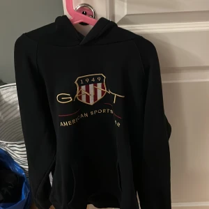 Svart hoodie från Gant med tryck - Svart hoodie från Gant med broderad logga och texten 'American Sportswear' framtill. Tröjan har huva och känguruficka, samt ett snyggt emblem i rött, vitt och guld. Perfekt för en sportig och avslappnad stil. Stl 176 . Passar en small/medium 