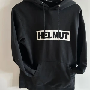 Helmut Lang Svart Hoodie - Svart hoodie från Helmut Lang med vit logga framtill. Klassisk modell med huva, snörning och känguruficka. Tillverkad i mjuk bomull och har långa ärmar. Perfekt för en avslappnad och stilren look.