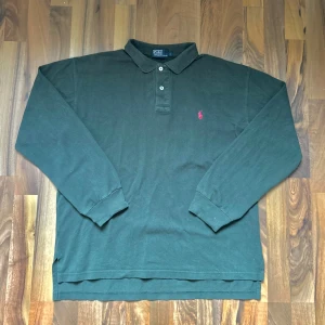 Mörkgrön pikétröja från Polo Ralph Lauren - Klassisk mörkgrön långärmad pikétröja från Polo Ralph Lauren med krage, knappar och ikonisk röd logga broderad på bröstet. Tillverkad i mjuk bomull och perfekt för en stilren och avslappnad look.