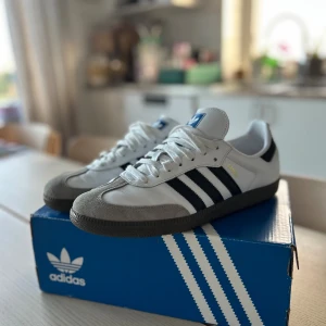 Adidas Samba vita och svarta sneakers - Adidas Samba sneakers i vitt skinn med svarta detaljer och klassiska tre ränder på sidan. Grå mockadetaljer vid tån och svart sula. Guldtexten 'SAMBA' på sidan ger en ikonisk touch. Perfekta för dig som gillar retrostil och streetwear.