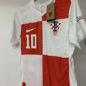 Kroatien Modric #10 Nike fotbollströja - Säljer en officiell Kroatien fotbollströja från Nike med Modric och nummer 10 på ryggen. Tröjan har rödvita blockrutor, Kroatiens landslagsemblem, korta ärmar och turneringspatch på ärmen. Materialet är lätt och ventilerande polyester, perfekt för match eller träning.