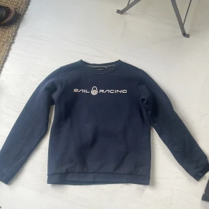 Mörkblå Sail Racing sweatshirt - Mörkblå sweatshirt från Sail Racing med vit logga framtill. Klassisk rund halsringning och långa ärmar. Mjuk insida och ribbade muddar vid ärmslut och nederkant. Perfekt för dig som gillar stilren och sportig look.