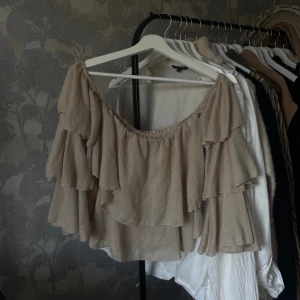 Beige offshoulder volangblus - Säljer en superfin beige blus med offshoulder och flera lager volanger. Toppen har vida ärmar med volangdetaljer och är croppad i modellen. 