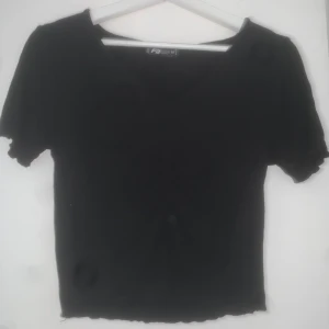 Svart ribbad topp  - Svart ribbad topp från FB Sister med v-ringning och korta ärmar. Modellen är croppad med vågig kant nertill och vid ärmslut. Liten scrunch i mitten av brösten. Enkel och stilren design som passar till många olika looks.