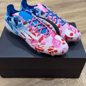 Adidas F50 x Bape fotbollsskor - Unika Adidas F50 x Bape fotbollsskor med färgstarkt camomönster i blått, rosa och rött. Skorna har snörning, platt sula med dobbar och cool hajdetalj på sidan. Ovandelen är i syntetmaterial och sulan skimrar i regnbågsfärger. Perfekta för dig som vill sticka ut på planen.