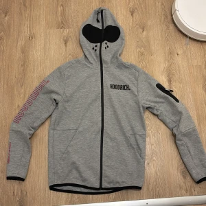 Grå hoodie och byxor från Hoodrich med dragkedja - Grå hoodie från Hoodrich med svart dragkedja, huva och coola tryckdetaljer på bröst, rygg och ärm. Huva med mesh och ventilationshål, samt fickor och en extra ficka med dragkedja på ärmen. Perfekt för dig som gillar streetwear och snygga detaljer. Byxorna ingår 