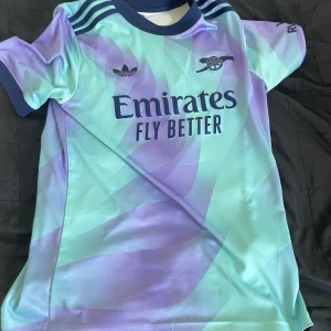 Arsenal x Adidas kortärmad fotbollströja - Snygg Arsenal fotbollströja från Adidas med korta ärmar och coolt mönster i ljusblått och lila. Tröjan har mörkblå detaljer vid krage och axlar, samt tryck med Emirates Fly Better på bröstet. Perfekt för dig som vill sticka ut på planen.