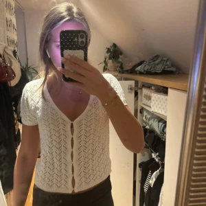 Vit stickad blus från Gina - Supersöt vit stickad blus från Gina med korta ärmar och v-ringning. Blusen har ett fint hålmönster, knappar framtill och är croppad i modellen. Perfekt för vår och sommar när du vill ha en luftig och trendig look.