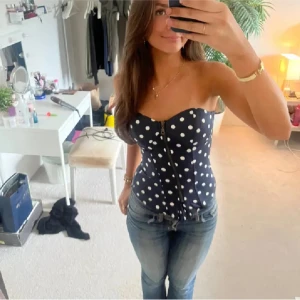 Mörkblå prickig korsett med dragkedja - Supersnygg mörkblå korsett med vita prickar och dragkedja framtill. Axelbandslös modell med markerad midja och lätt vadderade kupor. Perfekt att styla med jeans eller kjol för en trendig look.
