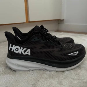 Svarta Hoka Clifton 9 sneakers - Säljer ett par svarta Hoka Clifton 9 sneakers med vit och svart tjock sula. Skorna har snörning, mesh-ovandel och stora vita HOKA-loggor på sidan. Perfekta för dig som gillar löpning eller vill ha riktigt bekväma sneakers till vardags. Använda 3-4 gånger, mycket bra skick 