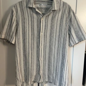 Randig kortärmad skjorta från Zara - Säljer en snygg kortärmad skjorta från Zara i storlek M. Skjortan är vit med svarta ränder, har knappar framtill och en avslappnad passform. Materialet är bomull och känns luftigt, perfekt för varmare dagar.