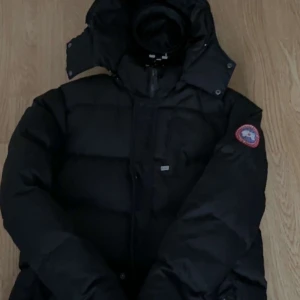 Svart dunjacka från Canada Goose - Säljer en svart dunjacka från Canada Goose med huva och klassisk logga på ärmen. Jackan har en puffad design, dragkedja framtill och flera fickor. Perfekt för kalla dagar och har en riktigt schysst streetstil.