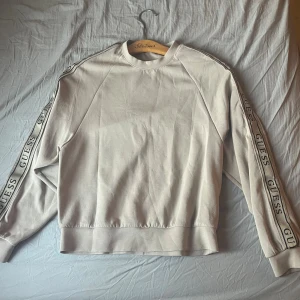 Beige sweatshirt från Guess - Snygg beige tunn sweatshirt från Guess med logotypband längs båda ärmarna. Tröjan har rund hals och långa ärmar med ribbade muddar. 