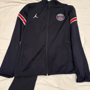 PSG x Jordan svart träningsdress - Svart träningsjacka från PSG och Jordan med dragkedja, lång ärm och broderad klubbmärke på bröstet. Röd och vit rand på ärmarna ger en sportig vibe. Perfekt för fotboll eller chill. Tillverkad i mjuk polyester för skön passform. Byxor ingår!!
