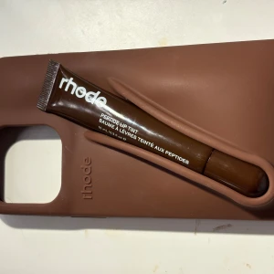Rhode Phone Case med Lip Tint-fack - Unik Rhode mobilskal i brunt silikon med integrerat fack för Rhode Peptide Lip Tint. Skalet är i gott skick och skyddar din telefon samtidigt som du alltid har läppbalsamet nära till hands. Passar utmärkt för dig som vill kombinera stil och funktion.
