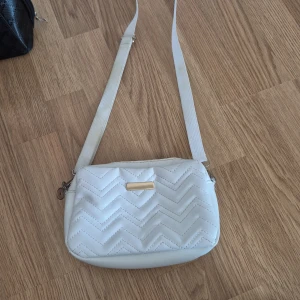 Vit quiltad axelväska med zigzag-mönster - Snygg vit axelväska med quiltat zigzag-mönster och justerbar rem. Väskan har en stilren rektangulär form och en liten guldfärgad detalj framtill. Perfekt för dig som vill ha en clean och trendig look.