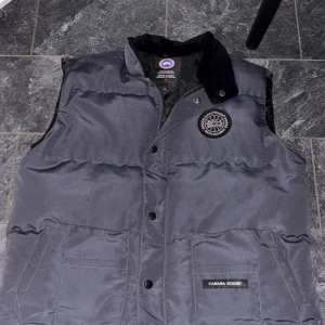 Svart dunväst från Canada Goose M - Säljer en svart dunväst från Canada Goose i storlek M. Västen har klassisk quiltad design, hög krage i mjukt material och två stora fickor framtill. Märkets logga sitter på bröstet och det finns både dragkedja och knappar framtill.