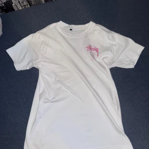Vit Stüssy t-shirt med rosa tryck - Vit t-shirt från Stüssy med klassiskt rosa logotryck på bröstet och större tryck på ryggen med städer som New York, Los Angeles, Tokyo, London och Paris. T-shirten har rund hals och korta ärmar, perfekt för en streetwear-look.