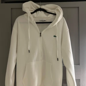 Vit hoodie från Lacoste med dragkedja - Vit hoodie från Lacoste i regular fit med klassisk krokodillogga broderad på bröstet. Tröjan har huva med snörning, dragkedja framtill och stora fickor. Mjuk bomullskänsla och långa ärmar gör den perfekt för chill dagar. Inga defekter 