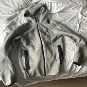 Nike tech  - Snygg ljusgrå hoodie från Nike med svart dragkedja och svarta detaljer vid fickorna. Tröjan har huva och Nike-logga på bröstet. Perfekt för chill eller träning.