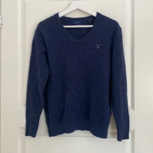 Mörkblå ulltröja med v-ringning GANT - Klassisk mörkblå stickad tröja från GANT i extra fin lammull. Tröjan har v-ringad hals, långa ärmar och diskret broderad logga på bröstet. Perfekt för lager-på-lager och snygg till både jeans och chinos.