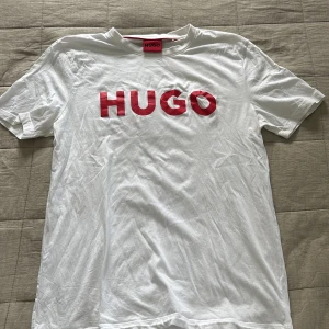 T-Shirt från Hugo - Säljer en vit t-shirt från Hugo med stor röd HUGO-logga på bröstet. Klassisk rund hals och korta ärmar. T-shirten är gjord i mjuk bomull och har en clean, enkel look som passar till jeans eller shorts.