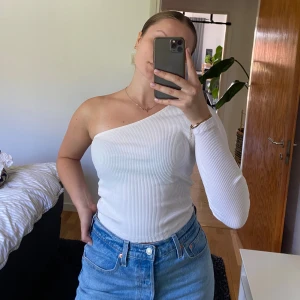 Vit ribbad oneshoulder-top - Säljer en vit ribbad topp med oneshoulder-design och lång ärm på ena sidan. Toppen är tight och har en stilren, minimalistisk look som passar perfekt till jeans eller kjol. Materialet är mjukt och stretchigt för en bekväm passform.